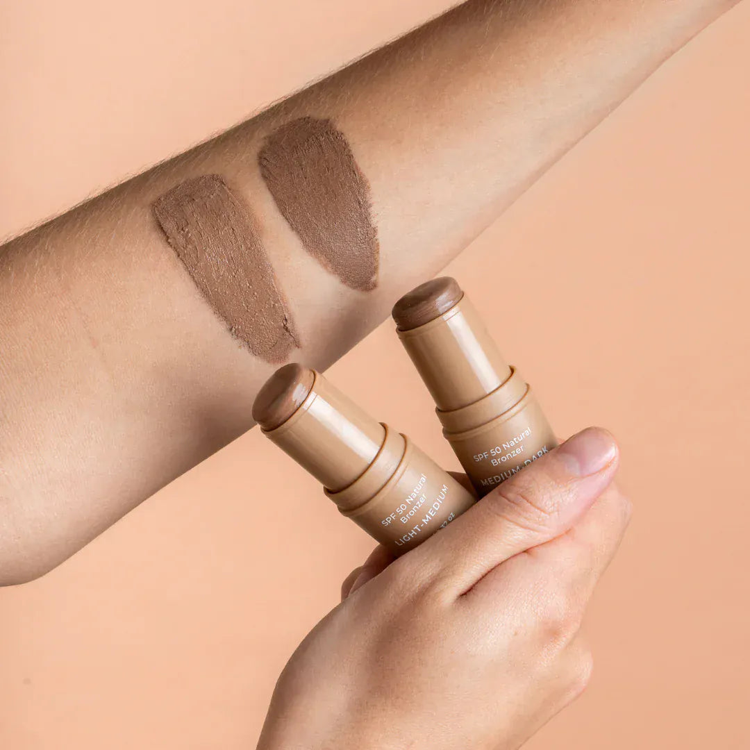 Person holding Avocado Zinc SPF 50 Natural Bronzer sticks with swatches on arm showing warm, bronzed shades.