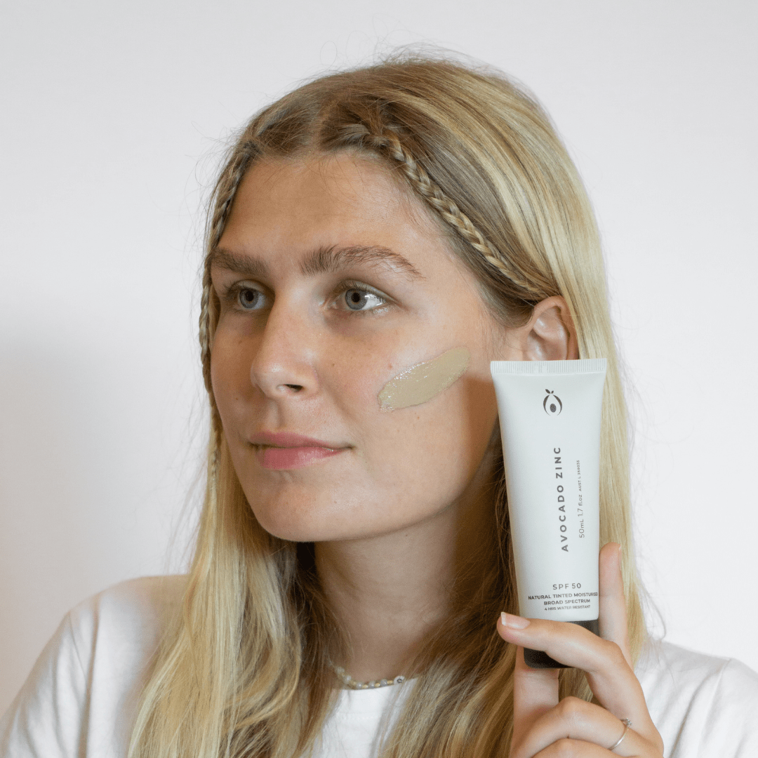 Woman applying Avocado Zinc SPF 50 Tinted Moisturiser on face for lightweight coverage and UV protection.