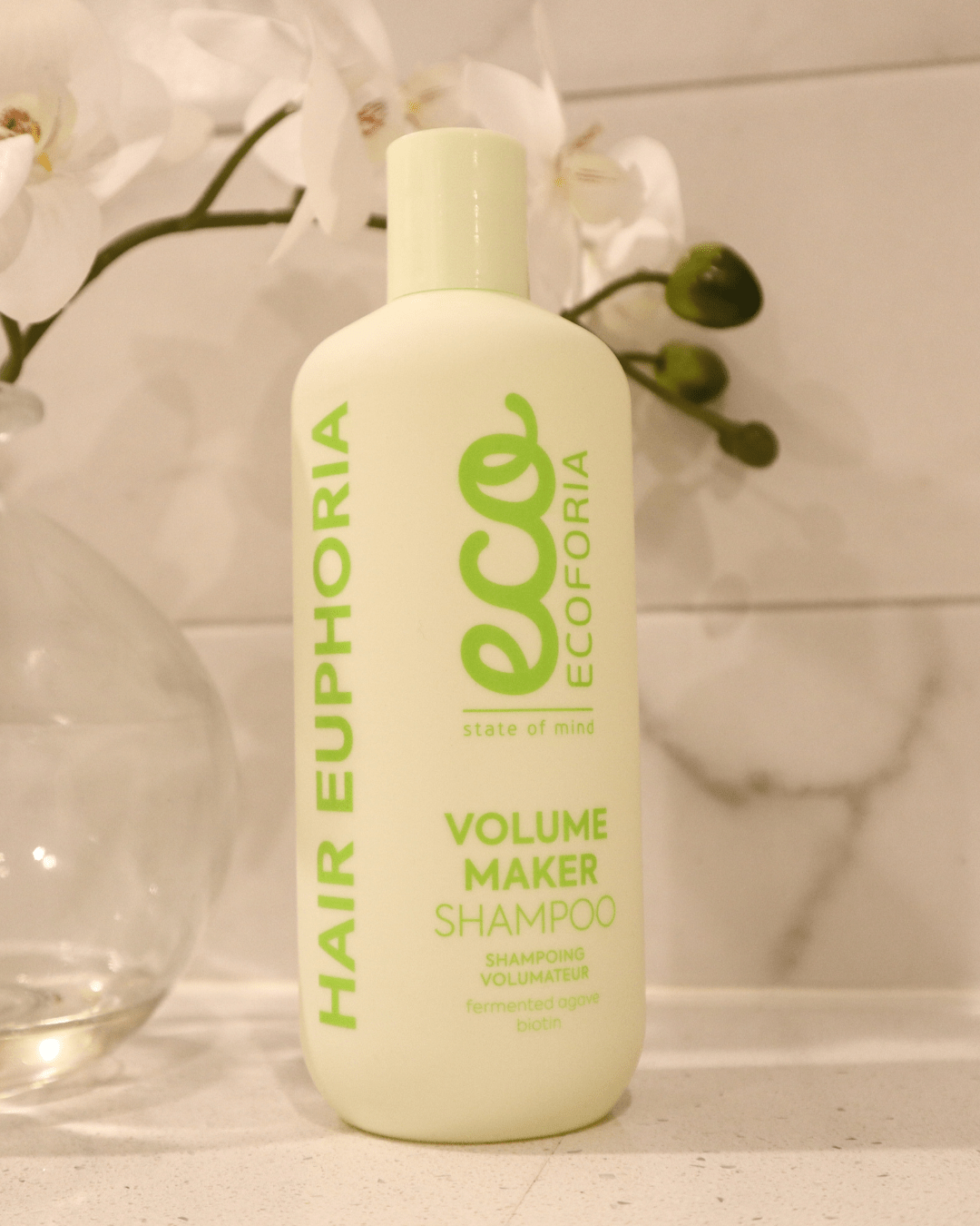 Bottle of Ecoforia Volume Maker Shampoo (400ml) for fuller, balanced hair, displayed on a bathroom shelf with orchids.