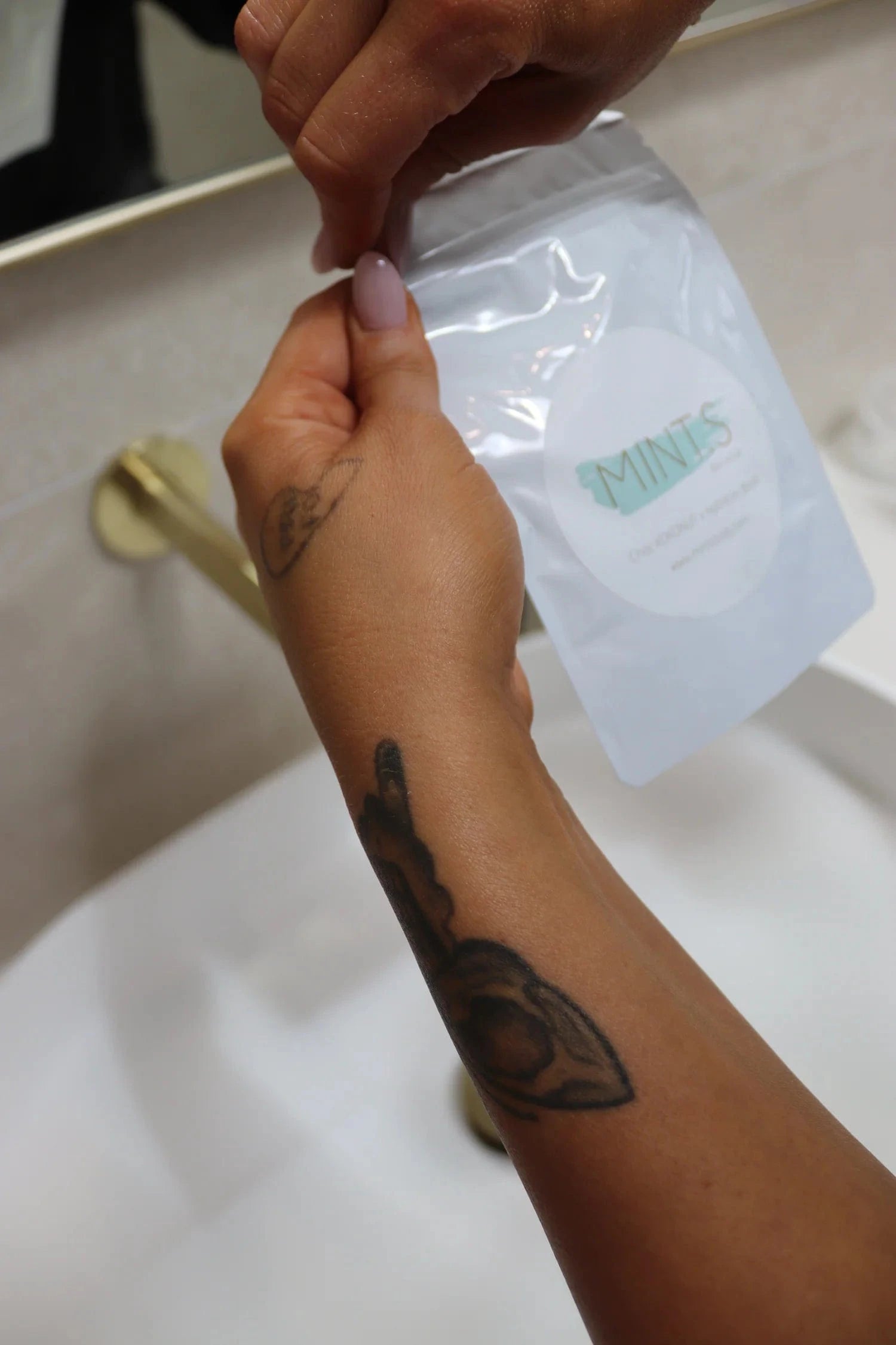 Person holding a package of Mint.s Face Scrub near a bathroom sink, showcasing the product's gentle exfoliation.