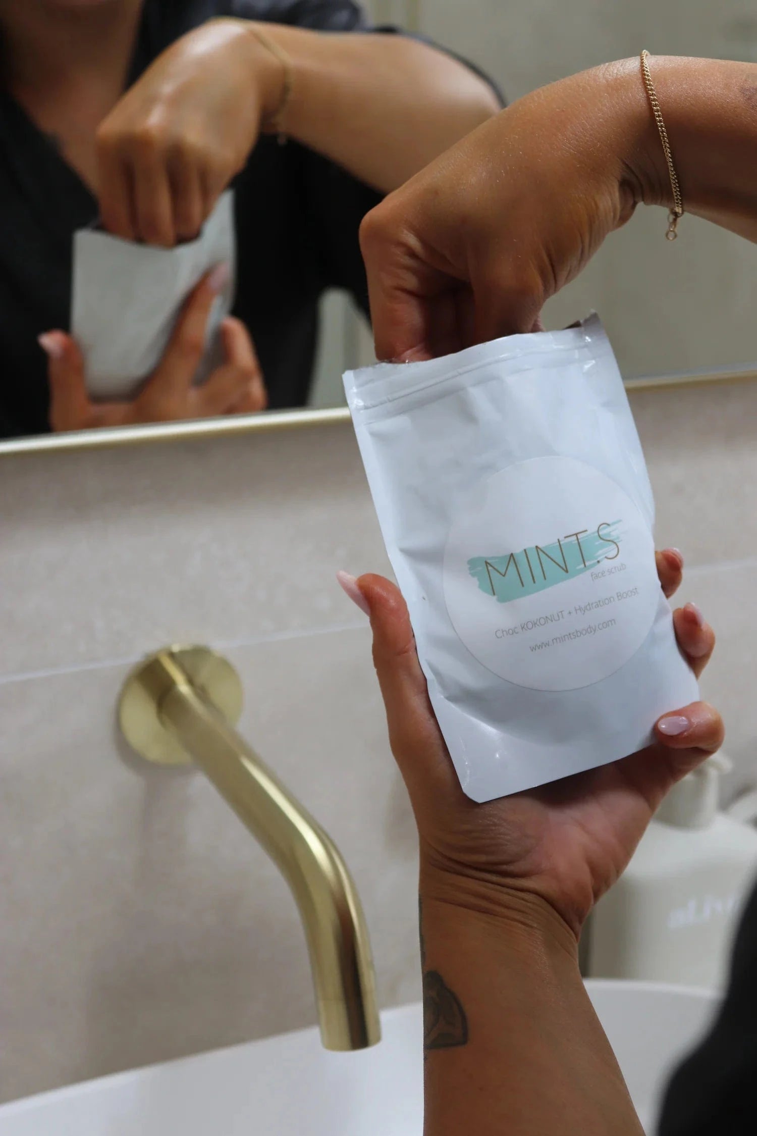 Person holding Mint.s Face Scrub packaging in front of a bathroom mirror, showcasing the product for gentle exfoliation.