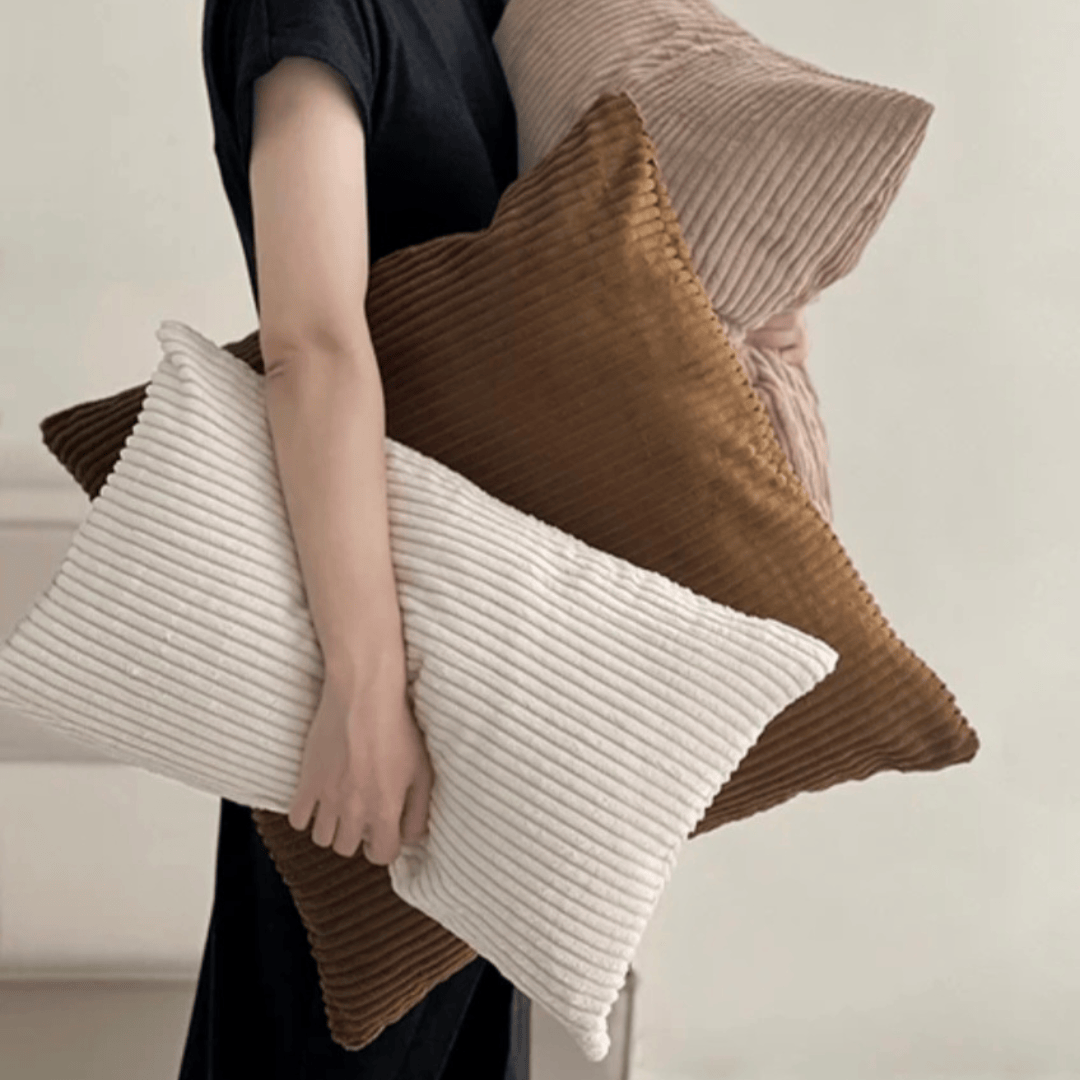 Person holding three textured cushions, including a Corduroy Cushion Cover Truffle in a warm earthy tone.