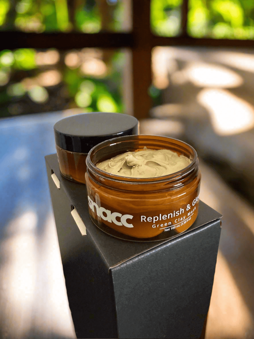 SNACC Beauty Replenish & Glow Green Clay Face Mask jar open on a stand with blurred natural background