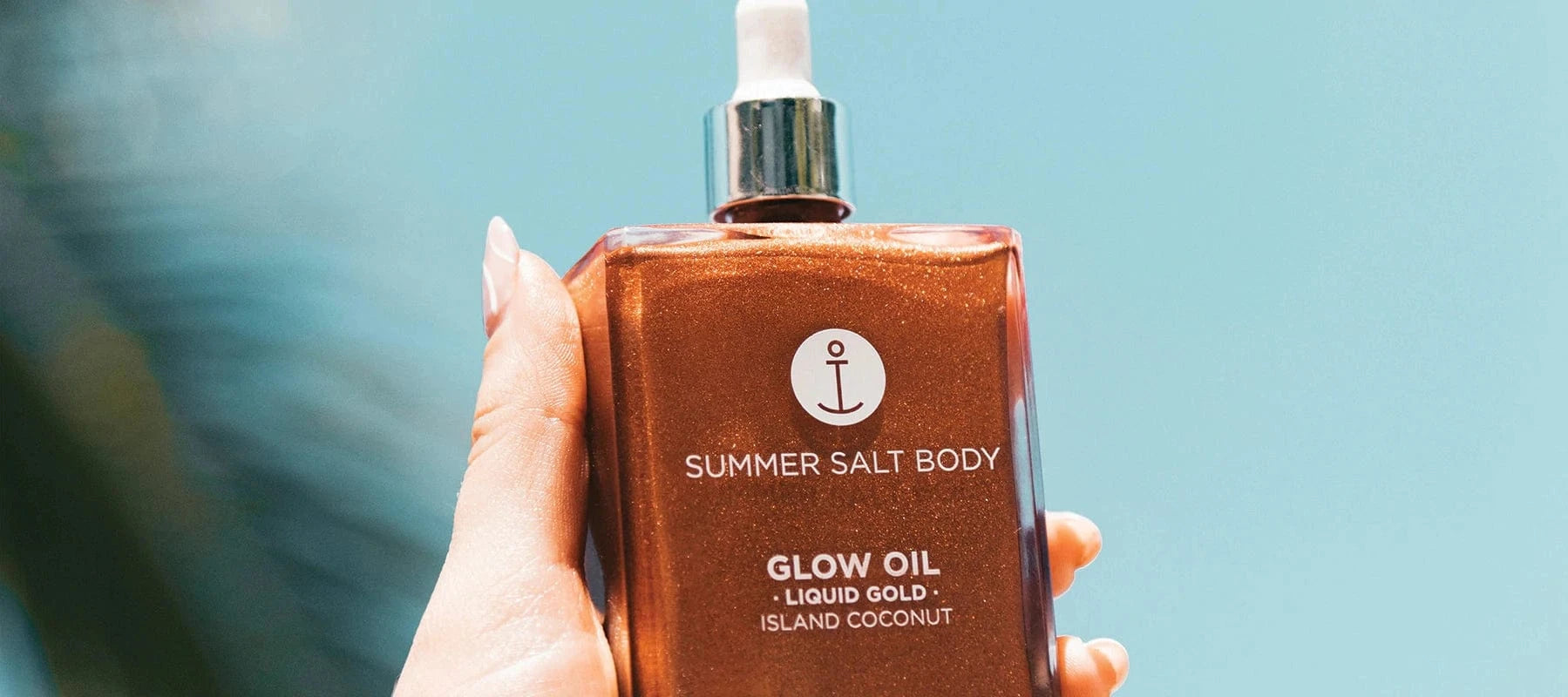 Summer Salt Body Liquid Gold Body Glow Oil bottle held against a blue sky background.