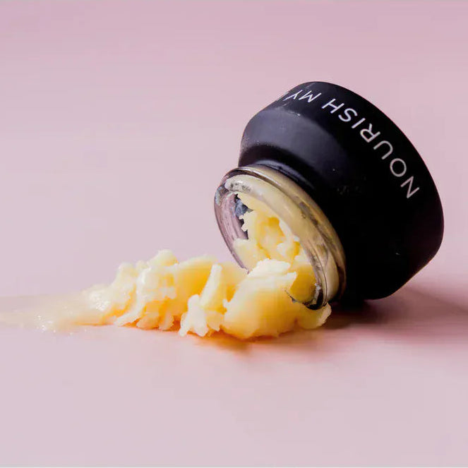 Nourish My Lips Lip Balm 15ml hydrating balm spilled from a black container onto a pink surface, showcasing its creamy texture for dry lips.