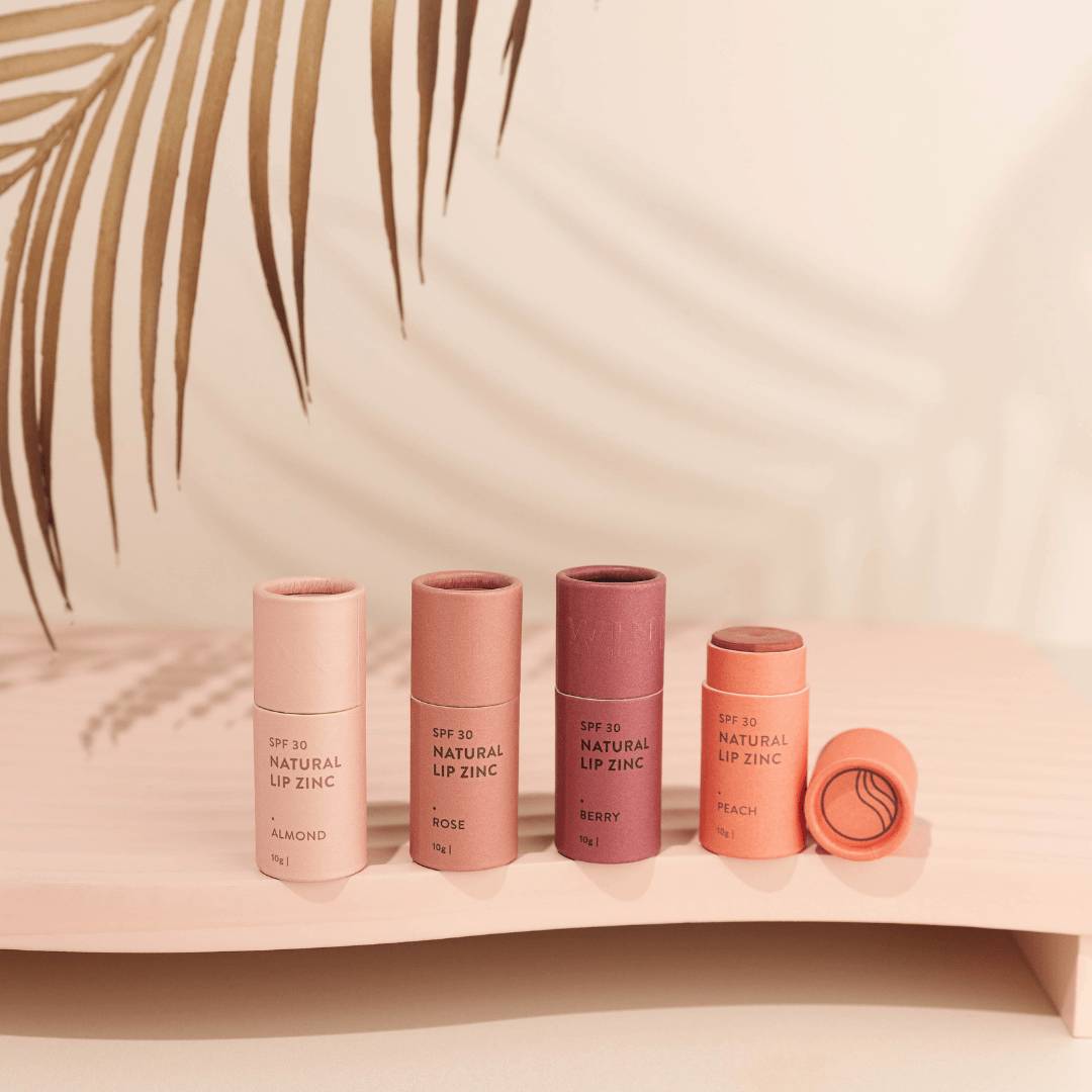 Four variations of Winki Zinc Tinted Natural Lip Zinc displayed on a pink surface with a palm leaf shadow in the background.