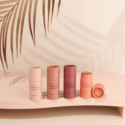 Four variations of Winki Zinc Tinted Natural Lip Zinc displayed on a pink surface with a palm leaf shadow in the background.